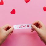 Hands holding an 'I Love U' note on a pink background with red hearts. Perfect for Valentine's themes.